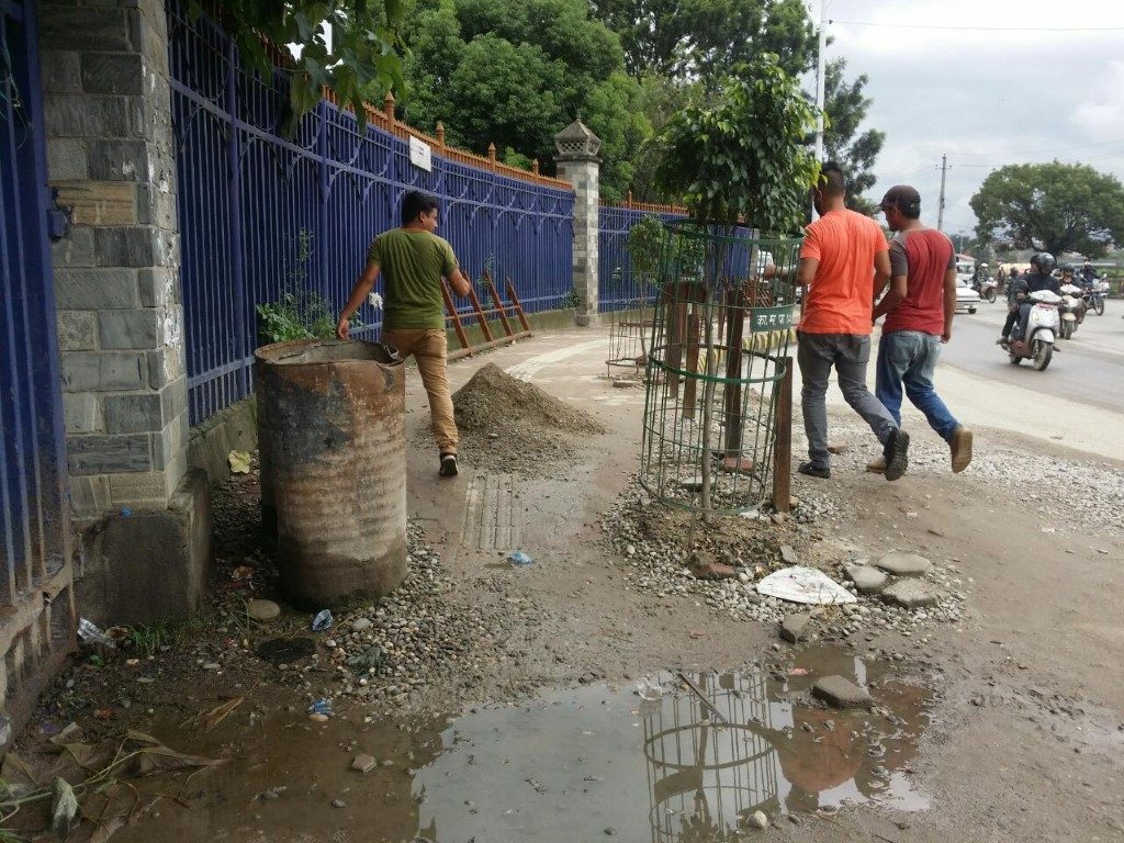 अव्यस्थित सडकपेटीको तस्बिर। भूईँभरी हिलो छ र सडकपेटिको बिचमा नै रूखको बिरूवा समेत रहेको छ। दुईजना मान्छेहरू अफ्टेरो गरेर पेटीमा हिड्दैछन्।