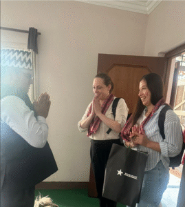 A warm and friendly meeting. Two women are standing inside a room, facing a man who is greeting them with his hands pressed together in a traditional gesture of respect, often seen in South Asian cultures. The women are smiling and returning the gesture. Both women are wearing red patterned scarves around their necks and have backpacks. One of them is holding a black gift bag with the word "MYRIGHT" and a star logo printed on it. The room has a wooden door and a window with curtains. The atmosphere appears cordial and welcoming.
