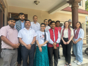 A group of twelve people is standing together outside a building. They are posing for a group photo. The group consists of both men and women, with a mix of casual and semi-formal attire. Two women on the right are wearing white shirts with red patterned scarves around their necks. One woman in the front center is wearing a white t-shirt with a printed image and text, paired with a teal skirt. The man next to her is wearing a red vest over a white shirt. The background shows a beige building with a red-tiled roof and a decorative column. The atmosphere appears to be friendly and relaxed.