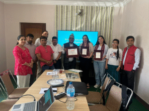 A group photo of Ten people in a room, with a mix of men and women standing together. In the center, three people are holding framed certificates. The man in the middle is wearing a traditional Nepali hat and a suit, while the two women on either side of him are dressed in casual business attire. The woman on the left is wearing a scarf. Behind them, a large screen displays the text "Inclusive Education and Human Rights Project" The room has a table in the foreground with a laptop, a water bottle, glasses, and some papers. The walls have a patterned wallpaper, and there are several chairs around the table.