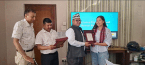 A group of four people in a room, with two men on the left and a man and a woman on the right. The man in the center is wearing a traditional Nepali hat and is handing a framed certificate to the woman, who is smiling and wearing a striped shirt with a red scarf. The two men on the left are observing the exchange, with one holding a similar framed item. In the background, there is a television screen displaying the text "Inclusive Education and Human Right Project," and a logo with the word "MYRIGHT." The room has striped wallpaper, and there is a wooden table with a helmet and some books on it.