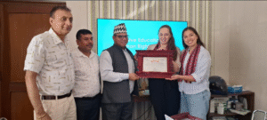 A group of five people is standing in a room, posing for a photo. The group consists of three men and two women. The man in the center is wearing a traditional Nepali hat and is holding a framed certificate with the two women. The woman on the right is wearing a striped shirt and a red patterned scarf, while the woman next to her is wearing a black top. The men on the left are dressed in light-colored shirts, with one wearing a patterned shirt and the other a plain one. Behind them, there is a television screen displaying the words "Inclusive Education and Human Rights Project." The room has patterned wallpaper and a wooden door on the left. There are some books and a helmet on a shelf in the background.