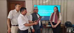 A group of four people is standing in a room. Three men and one woman are present. The man on the left is wearing a light-colored short-sleeve shirt with a pattern and beige pants. The second man is wearing a white short-sleeve shirt and dark pants, holding a red book or folder. The third man, in the center, is wearing a traditional Nepali hat, a gray vest over a light blue shirt, and dark pants. The woman on the right is wearing a white blouse and a red patterned scarf around her neck. She has long brown hair. Behind them, there is a large screen displaying the text "Inclusive Education and Human Rights Project" with a blue background. The room has patterned wallpaper, and there is a wooden cabinet with a helmet and some books on it.