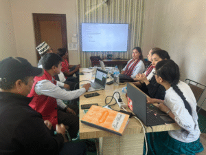 A group of people in a meeting room. There are eight individuals seated around a rectangular table, engaged in discussion. Most of them have laptops open in front of them. The room has a large flat-screen TV mounted on the wall displaying a presentation or document. The people are dressed in a mix of casual and traditional attire, with one person wearing a distinctive patterned hat. On the table, there are water bottles, a power strip with cables, and a couple of notebooks, one of which is labeled "Register." The atmosphere appears to be focused and collaborative.