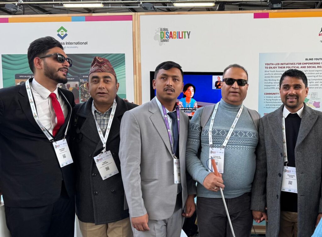 A group of five men standing together at an event. They are all wearing formal or semi-formal attire, with name badges hanging from lanyards around their necks. The man on the far left is wearing a black suit, white shirt, and red tie, and has sunglasses on. The second man is wearing a dark coat, plaid shirt, and a traditional Nepali hat. The man in the center is dressed in a light gray suit and has a purple lanyard. The fourth man is wearing a blue patterned sweater, sunglasses, and is holding a white cane, indicating he is visually impaired. The man on the far right is in a gray overcoat and black sweater. Behind them, there are informational posters and a screen displaying a woman and a sign language interpreter. The background signage includes the words "Global Disability Summit." The group appears to be at a conference or summit focused on disability rights and inclusion.