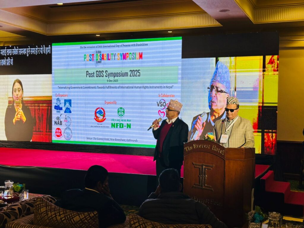 "Two male speakers wearing formal attire and Nepali topi stand at a podium and onstage during the ‘Post GDS Symposium 2025’ at The Everest Hotel, Kathmandu. Behind them, a large LED screen displays the event title, logos of co-organizers and collaborators, and a slide featuring the theme ‘Internalizing Government Commitments Towards Fulfilments of International Human Rights Instruments in Nepal.’ A sign language interpreter appears on the left side of the screen, and a large projected image of one speaker appears on the right. Audience members are seated in the foreground watching the session."