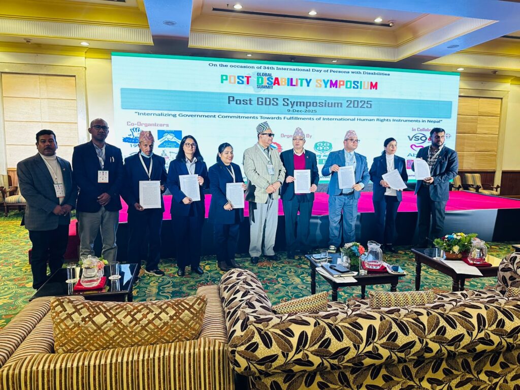 "A group of nine individuals stands in a row onstage during the ‘Post GDS Symposium 2025’ event at The Everest Hotel, Kathmandu. Each person is holding a document, posing for a formal group photo. They are dressed in business attire, with some wearing Nepali topi. Behind them, a large LED screen displays the event title, theme, and logos of co-organizers and collaborators. In the foreground, sofas, tables with water pitchers, and flower arrangements are visible, indicating a formal conference setup."