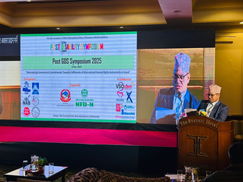 "An indoor conference hall setup with a large LED screen displaying Nepali text, sign language interpretation, and a digital timer showing 07:36. A man wearing a coat, tie, and traditional Nepali topi is standing at a podium, reading or presenting from a laptop. The stage has red carpeting, and the background includes wooden paneling and soft yellow lighting."