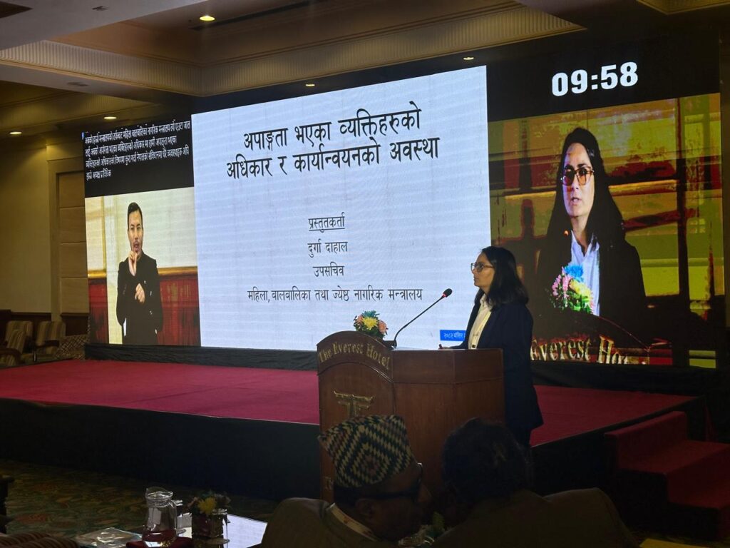 "A woman presenter stands at a podium labeled ‘The Everest Hotel,’ delivering a presentation in a conference hall. Behind her, a large LED screen displays a slide in Nepali titled ‘अपाङ्गता भएका व्यक्तिहरूको अधिकार र कार्यान्वयनको अवस्था,’ along with her name and designation. The screen also shows a sign language interpreter on the left and her live video feed on the right. A digital timer at the top right reads 09:58. Audience members sit in front, watching the session in a well-lit event setting."