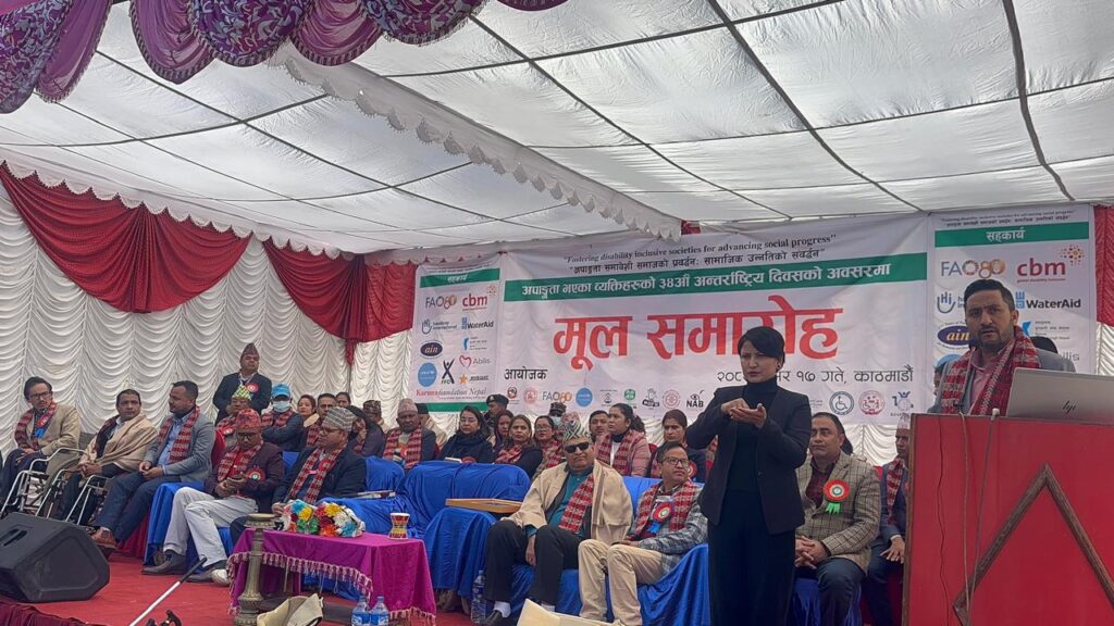A diverse group of people sit under a white tent with red drapes during an event. A speaker stands at a podium. A sign with logos hangs behind, conveying a formal, inclusive atmosphere.