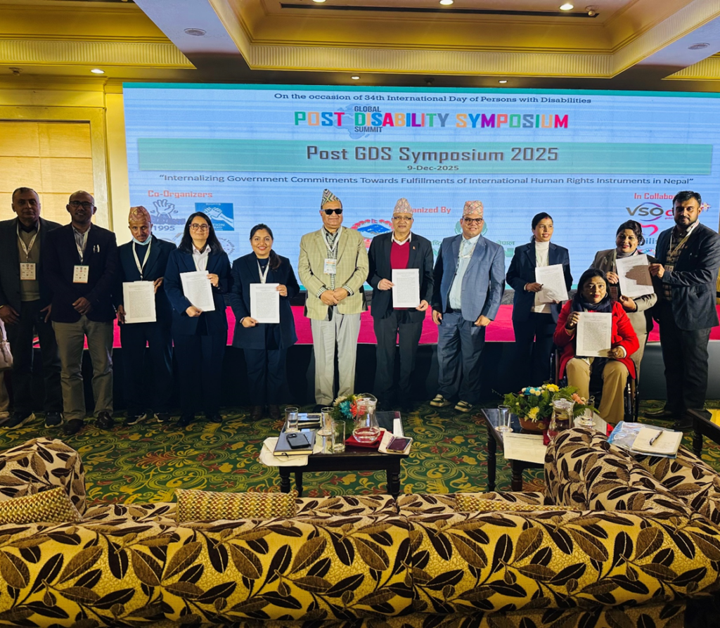 A group of men and women standing on a stage at a formal event, holding printed documents and posing for a group photo. Behind them, a large screen displays the title “Post GDS Symposium 2025” organized on the occasion of the 34th International Day of Persons with Disabilities. Participants are dressed in formal attire, including suits and traditional Nepali topi, and one woman is seated in a wheelchair at the front. The setting appears to be a hotel conference hall with stage lighting, banners, and event materials visible.