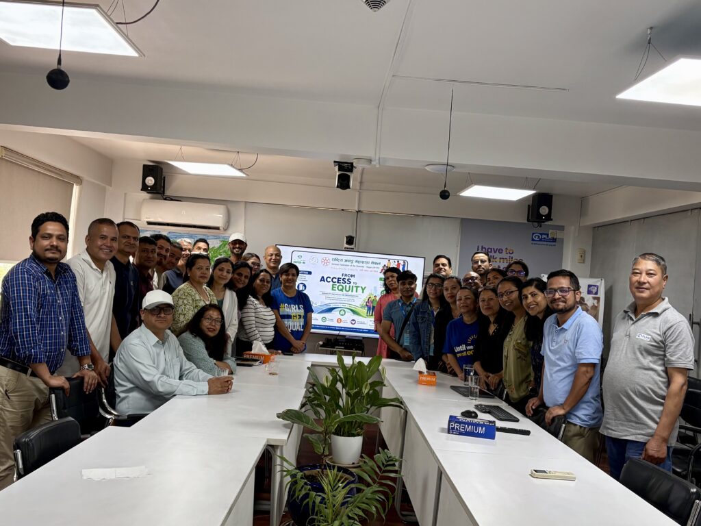 A large group of professional colleagues posing for a group photo in a well-lit meeting or conference room. The participants are diverse, consisting of both men and women, standing and sitting around a long, U-shaped white conference table. In the center foreground, potted green plants are arranged on the floor. In the background, a large digital display screen shows a presentation titled "FROM ACCESS TO EQUITY," featuring logos and text written in both English and Nepali script. The room has a modern office aesthetic with white walls, drop-ceiling panel lights, and audio-visual equipment.